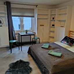 Pokój dzienny z łóżkiem, stolikiem z krzesłami i puszystym dywanikiem, przygotowany dla gości, z ręcznikami na łóżku i rośliną na stoliku.