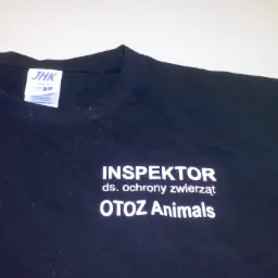 Czarna koszulka z białym nadrukiem 'INSPEKTOR ds. ochrony zwierząt OTOZ Animals', widoczna metka JHK, lekko zagnieciony materiał.