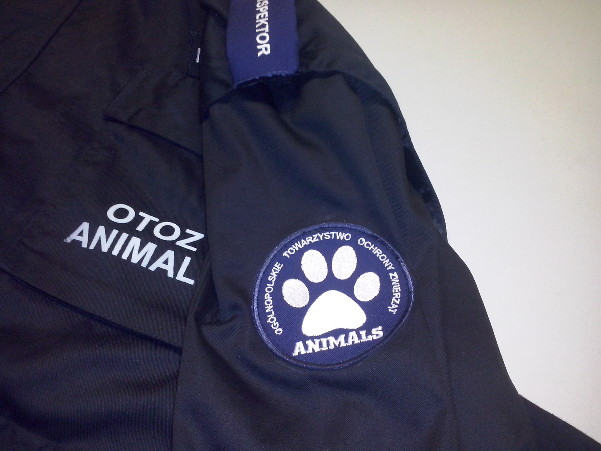 Fragment granatowej bluzy roboczej z naszywkami 'OTOZ Animals' i 'Inspektor', prezentujący detale haftu.
