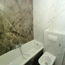 CENTRUM USŁUG - DANIEL NOŻEWSKI - Wykończona łazienka z wanną i podwieszaną toaletą, ściany wyłożone płytkami imitującymi marmur w odcieniach szarości i bieli, widoczny niebieski czyścik w wannie.