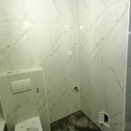 CENTRUM USŁUG - DANIEL NOŻEWSKI - Wykończona łazienka w Świnoujściu z płytkami gresowymi 60x120 cm imitującymi marmur na ścianach, podwieszany sedes z zabudową, widoczny fragment podłogi z ciemnymi płytkami i otwarty rewizyjny właz.