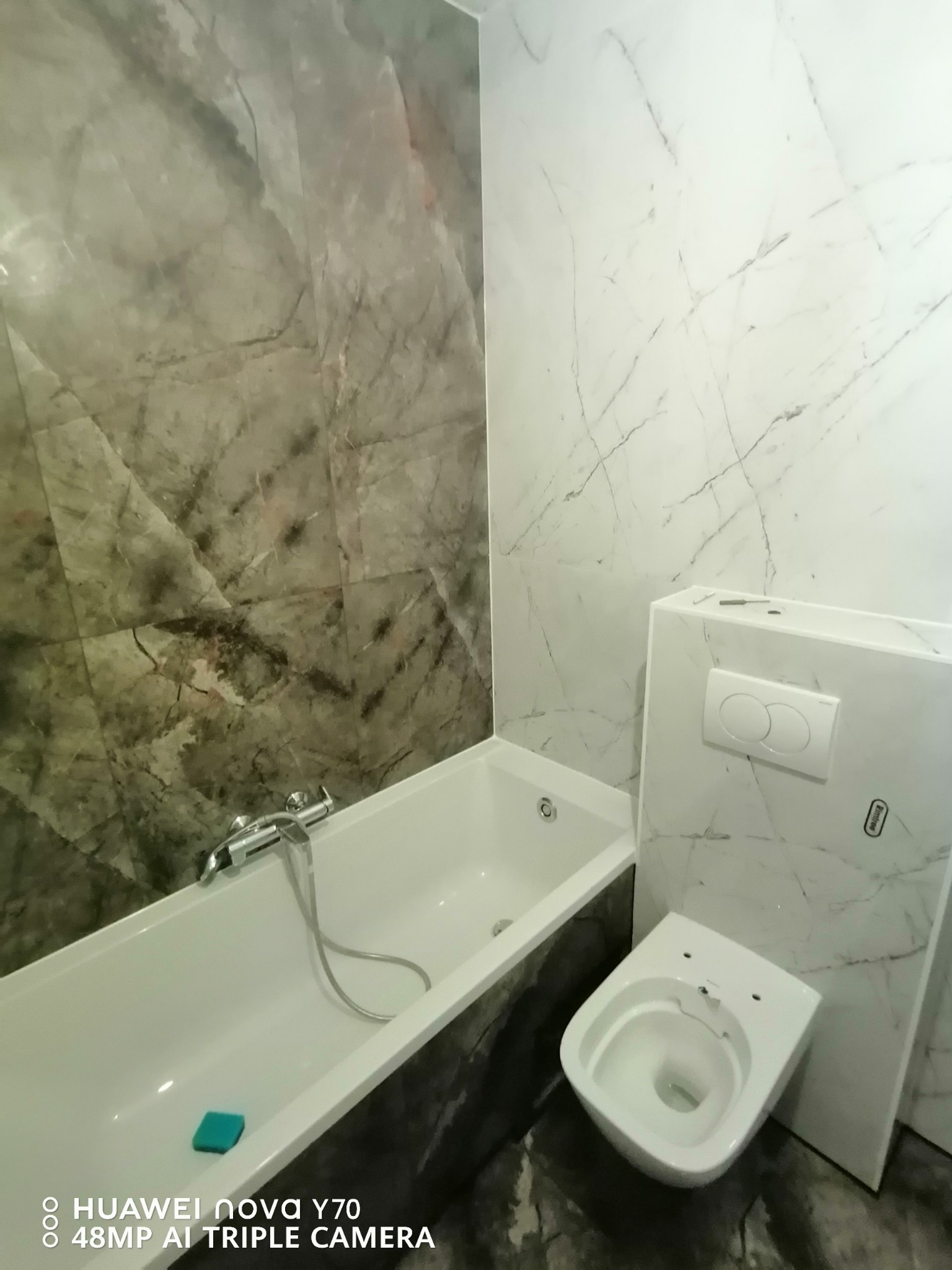 Wykończona łazienka z wanną i podwieszaną toaletą, ściany wyłożone płytkami imitującymi marmur w odcieniach szarości i bieli, widoczny niebieski czyścik w wannie.