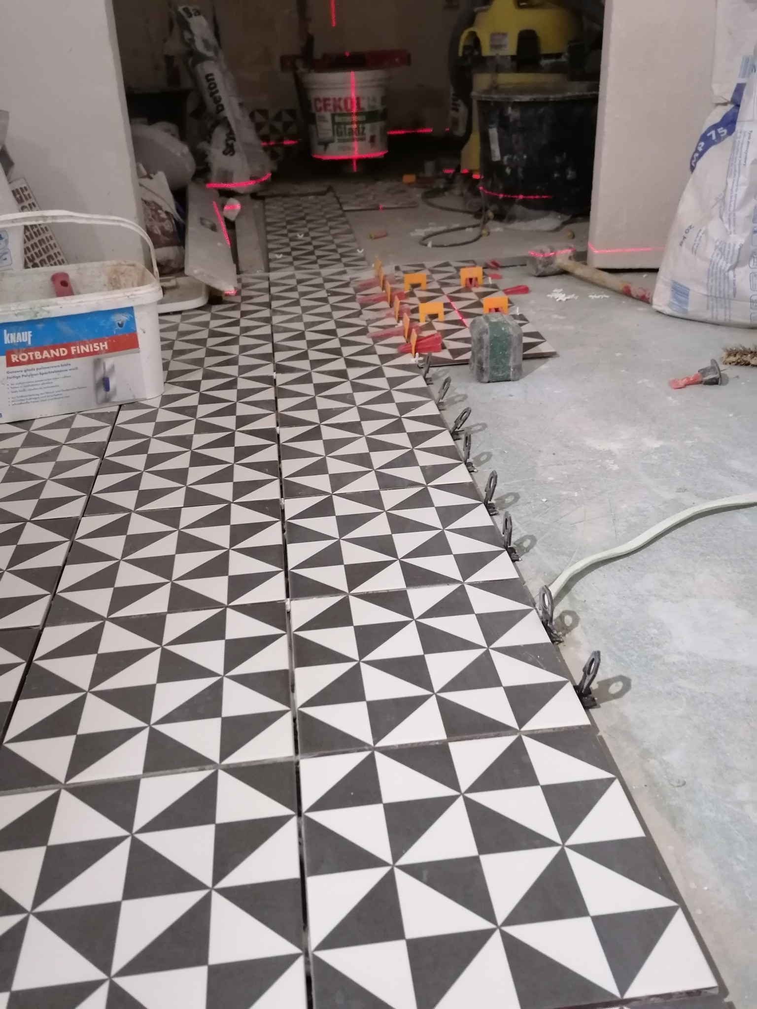 Układanie podłogi z czarno-białych płytek w geometryczny wzór, widoczne narzędzia glazurnicze, poziomica laserowa, worek z zaprawą oraz wiadro z masą szpachlową w tle, stan surowy.