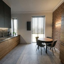 Projektowanie Wnętrz Katarzyna Ćwiek - Nowoczesna kuchnia z drewnianymi szafkami i czarnymi blatami, okrągły stół z krzesłami, ceglana ściana z oświetleniem. Minimalistyczny design, naturalne światło.