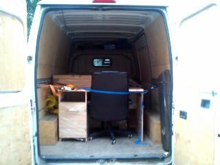 Wnętrze furgonetki z biurkiem, krzesłem i szafką zabezpieczonymi pasami. Widok z zewnątrz przez otwarte tylne drzwi, gotowe do transportu.