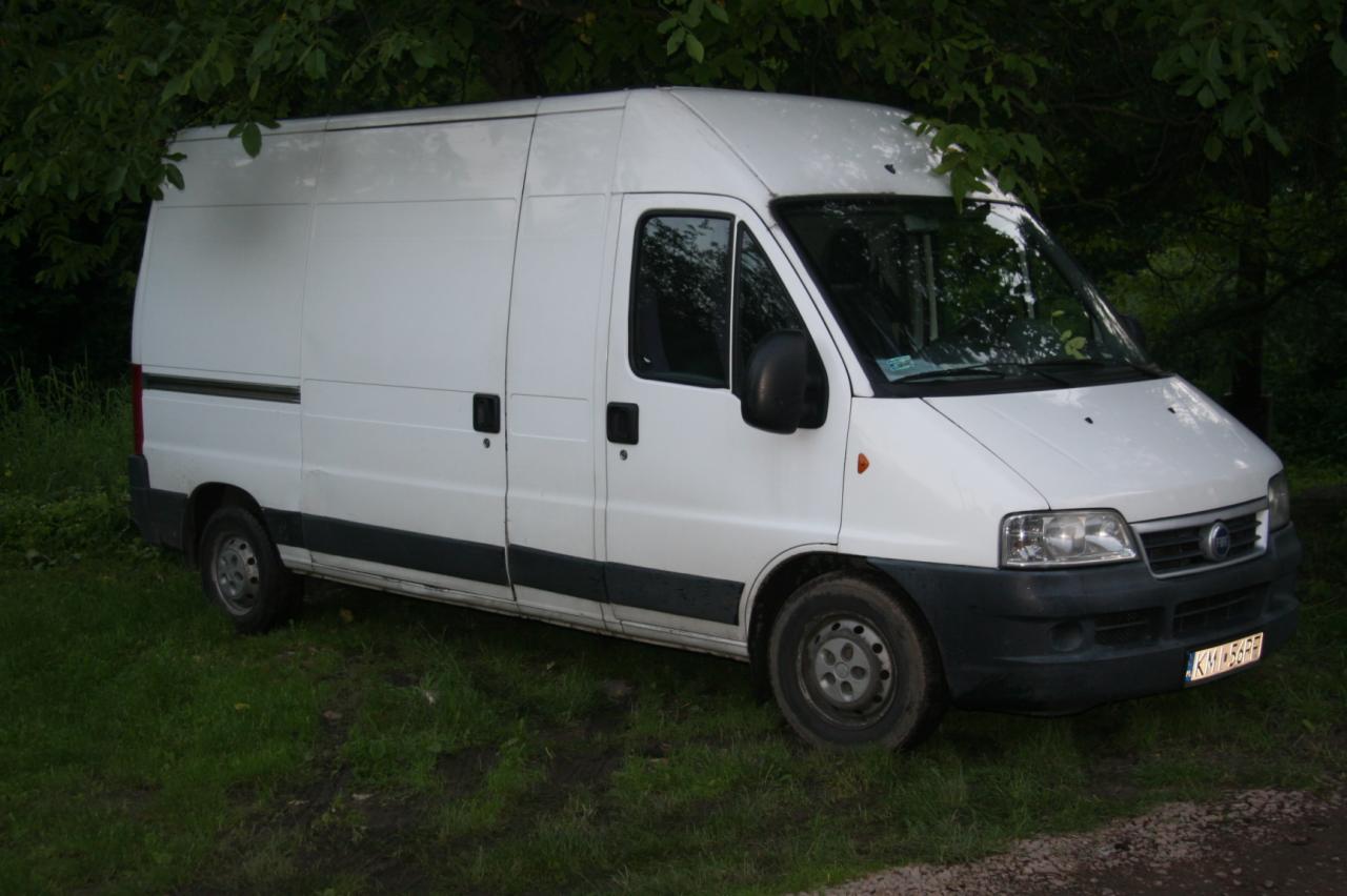 Biały samochód dostawczy Fiat Ducato na trawiastym terenie, częściowo zasłonięty przez gałęzie drzew. Widoczne tablice rejestracyjne.