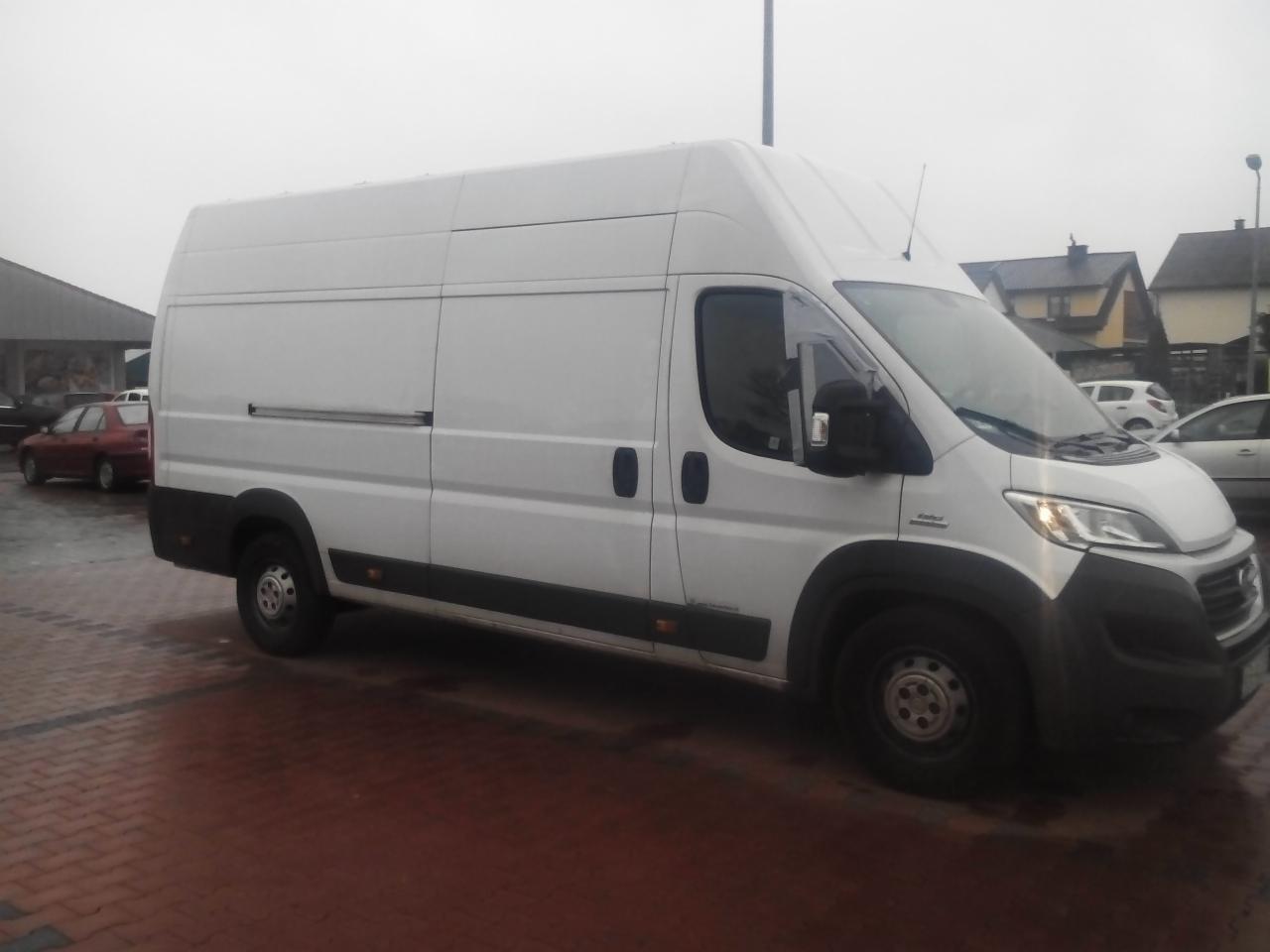 Biały furgon dostawczy Fiat Ducato z wysokim dachem, zaparkowany na kostce brukowej w pochmurny dzień. Widoczny z boku, zarys budynków w tle.