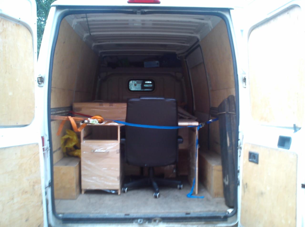 Wnętrze białego busa z otwartymi drzwiami, załadowane biurkiem, krzesłem i meblami zabezpieczonymi folią stretch i pasami transportowymi.