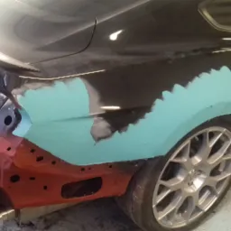 Czarny Mustang w trakcie naprawy blacharskiej, widoczne szpachlowane i zagruntowane elementy karoserii w kolorze turkusowym i czerwonym, brak zderzaka, srebrne felgi.