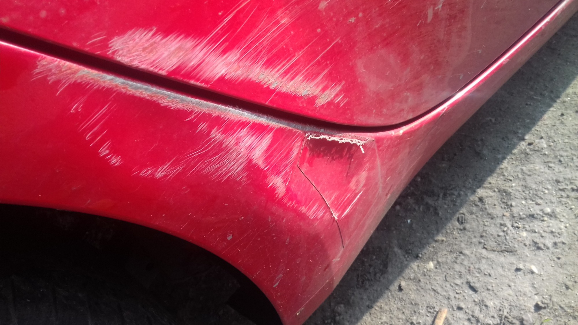 Czerwony błotnik samochodu Yaris z wyraźnymi zarysowaniami, odpryskami lakieru i wgnieceniem, widoczne ślady korozji.