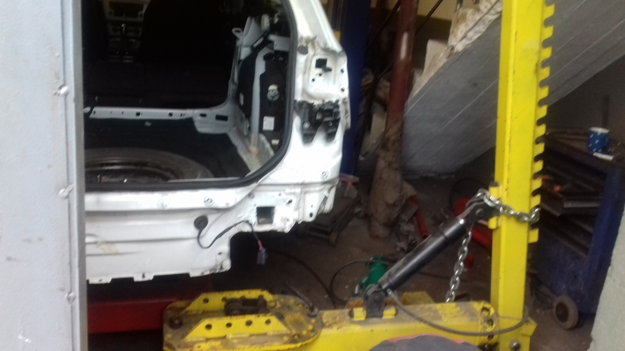 Biały samochód typu kombi, prawdopodobnie VW Passat, po uszkodzeniach tylnej części karoserii, wsparty na żółtym podnośniku w warsztacie.