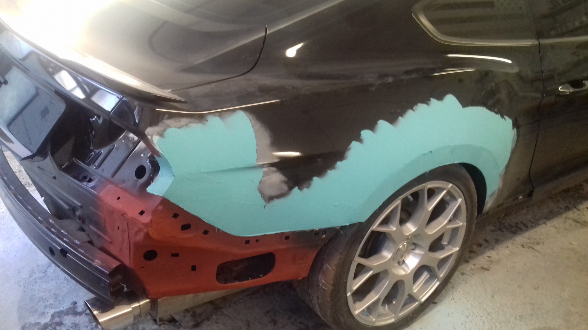 Czarny Mustang w trakcie naprawy blacharskiej, widoczne szpachlowane i zagruntowane elementy karoserii w kolorze turkusowym i czerwonym, brak zderzaka, srebrne felgi.