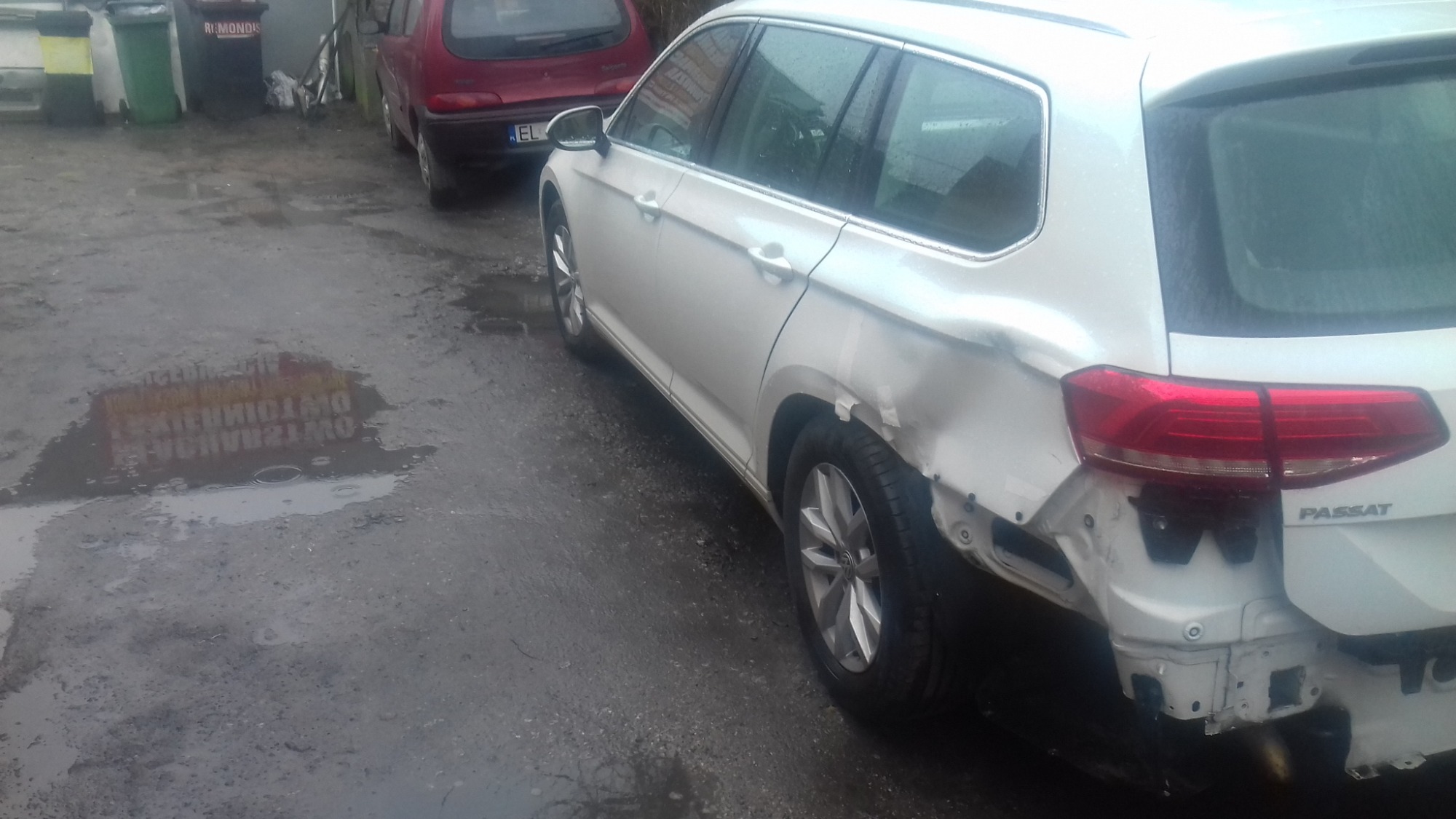Biały Volkswagen Passat Variant z uszkodzonym błotnikiem i zderzakiem, widoczne ślady uderzenia i brakujące elementy karoserii, zaparkowany na mokrym, szarym podłożu, w tle inne pojazdy i pojemniki...