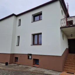 Dwukondygnacyjny dom z elewacją w kolorze białym i brązowym, z balkonem z czerwoną balustradą i schodami wejściowymi z brązowych płytek.