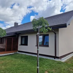 Jasny, otynkowany dom jednorodzinny z brązowymi detalami, widok z ogrodu w słoneczny dzień.