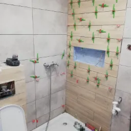 Układanie płytek w łazience z użyciem klipsów poziomujących. Widoczne fragmenty armatury i narzędzi. Częściowo wykończona ściana z płytek imitujących drewno.