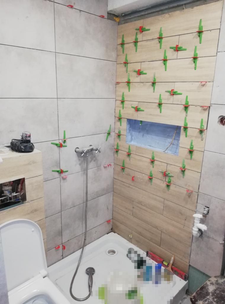 Układanie płytek w łazience z użyciem klipsów poziomujących. Widoczne fragmenty armatury i narzędzi. Częściowo wykończona ściana z płytek imitujących drewno.