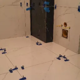 Łazienka w trakcie układania płytek: białe marmurowe płytki na ścianach i podłodze, czarne płytki w rogu, niebieskie klipsy poziomujące, widoczne rury instalacyjne.