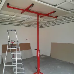 Montaż płyty gipsowo-kartonowej na suficie za pomocą podnośnika, widoczna drabina z wkrętarką, surowy stan wykończenia wnętrza.