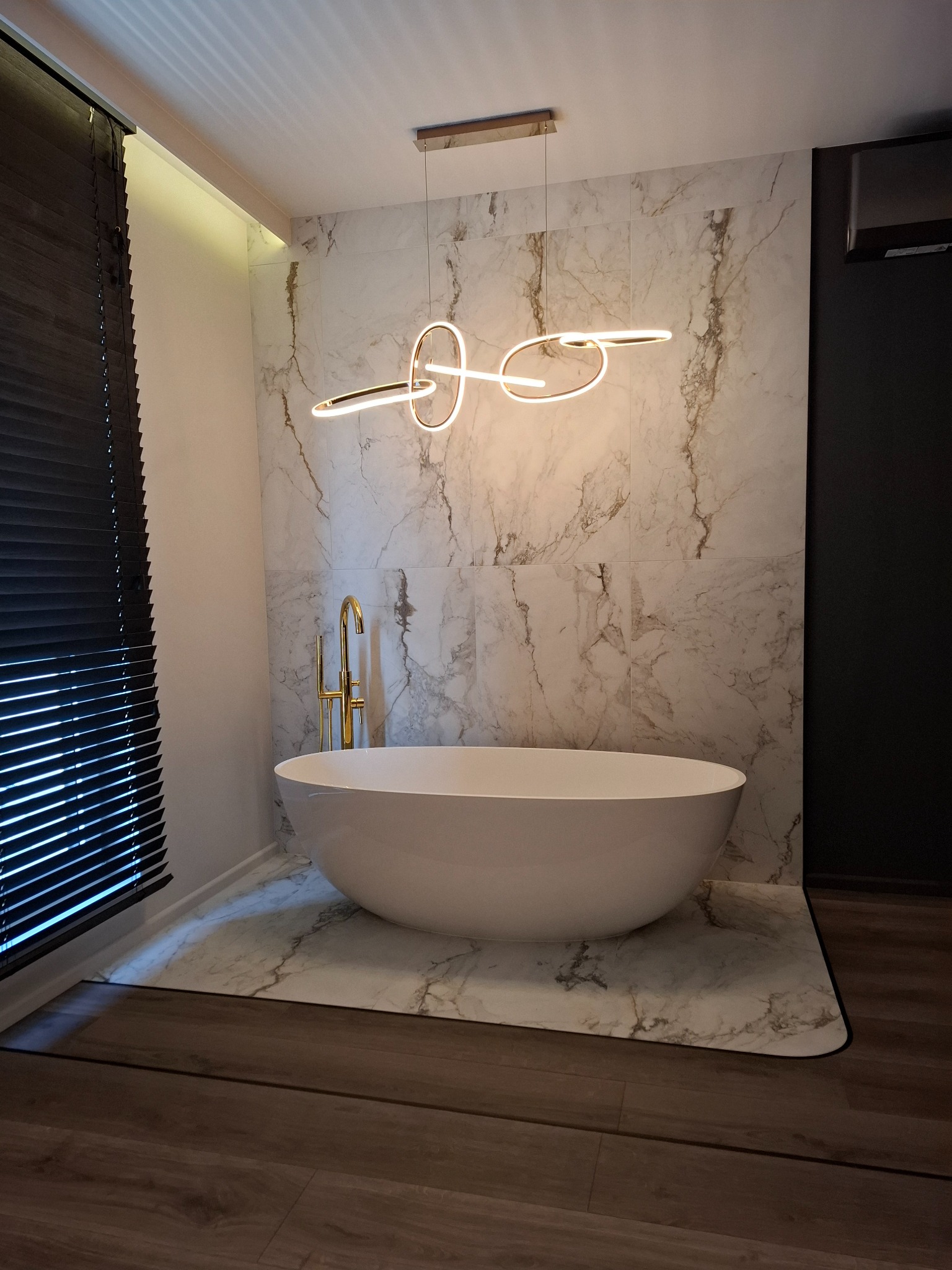 Nowoczesna łazienka z wolnostojącą wanną, złotą armaturą i marmurową ścianą. Unikalna lampa sufitowa dodaje elegancji. Widoczne żaluzje w oknie.