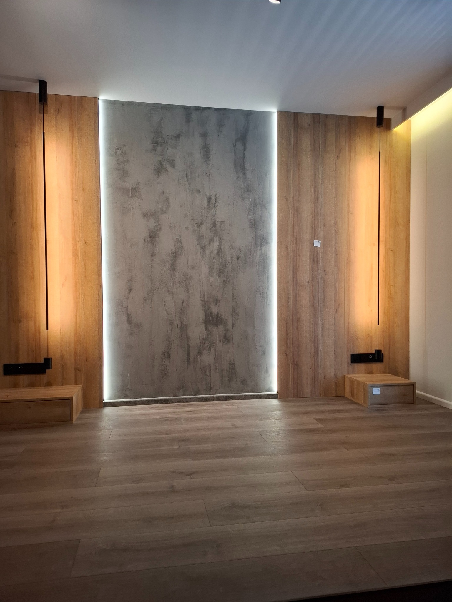 Nowoczesna sypialnia z drewnianą boazerią, betonową ścianą i podświetleniem LED. Minimalistyczne lampy wiszące i drewniana podłoga tworzą spójną całość.