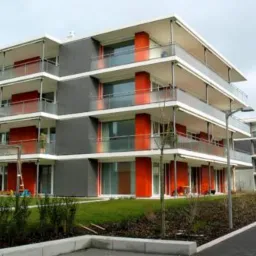 Nowoczesny, trzypiętrowy budynek mieszkalny z balkonami, szarą elewacją i pomarańczowymi akcentami, widok z zewnątrz na tle pochmurnego nieba.