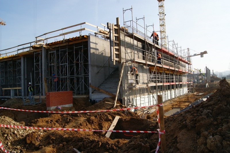 Wznoszenie konstrukcji żelbetowej budynku z użyciem szalunków, widoczne rusztowania i pracownicy w pomarańczowych kamizelkach, ziemia zabezpieczona taśmą ostrzegawczą.