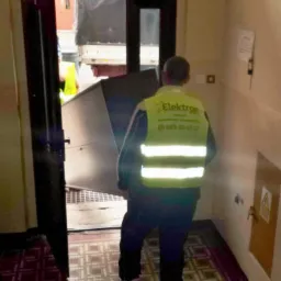 Pracownik w kamizelce firmy Elektron wnosi duży metalowy przedmiot do budynku, widoczny samochód dostawczy, przeprowadzka.