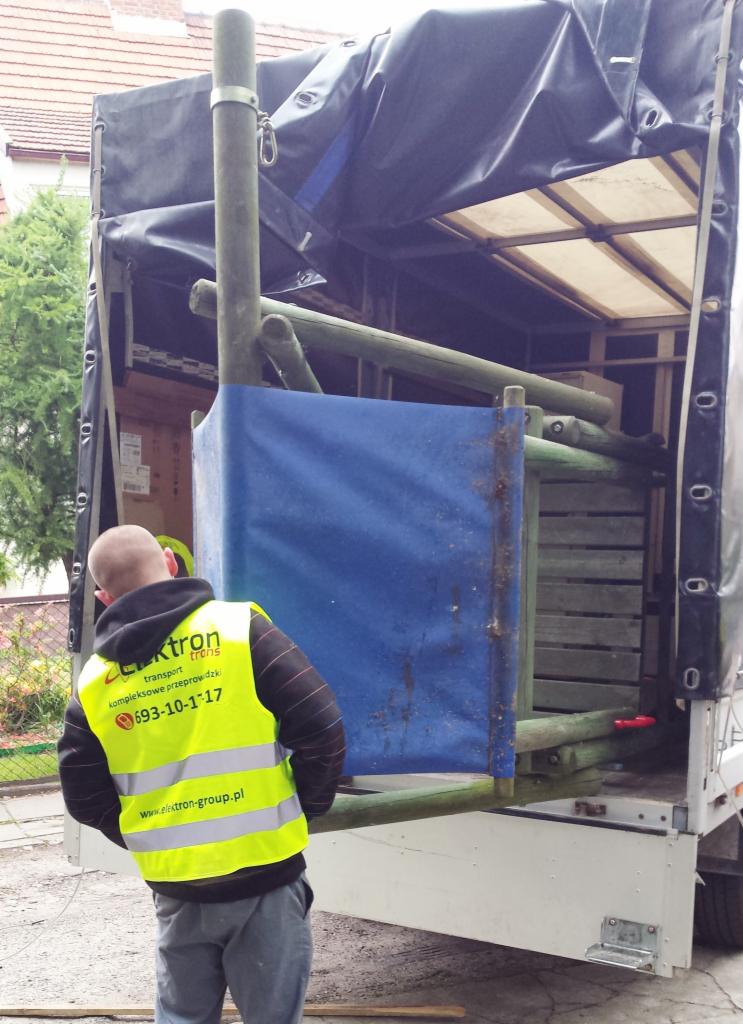 Mężczyzna w kamizelce firmy transportowej przy otwartej pace busa z drewnianą konstrukcją w środku, zabezpieczoną plandeką.