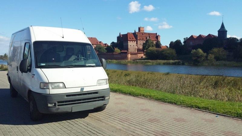 Biały bus transportowy na tle zamku w Malborku i rzeki Nogat. Pojazd zaparkowany na chodniku, błękitne niebo w tle. Dzień.