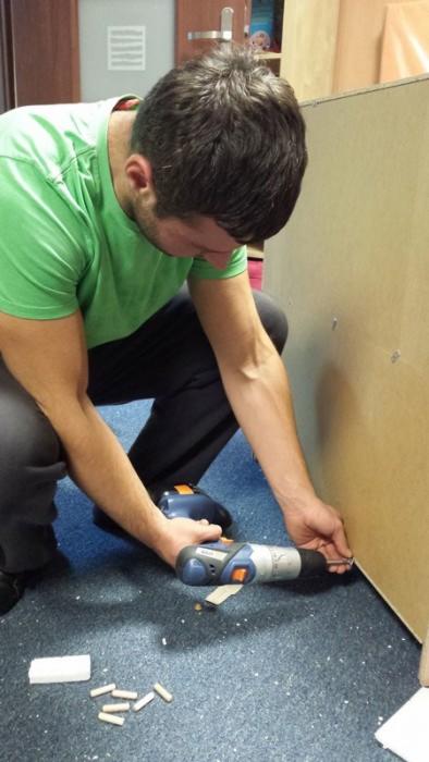 Mężczyzna w zielonej koszulce skręca płytę meblową wkrętarką. Na podłodze leżą kołki montażowe.