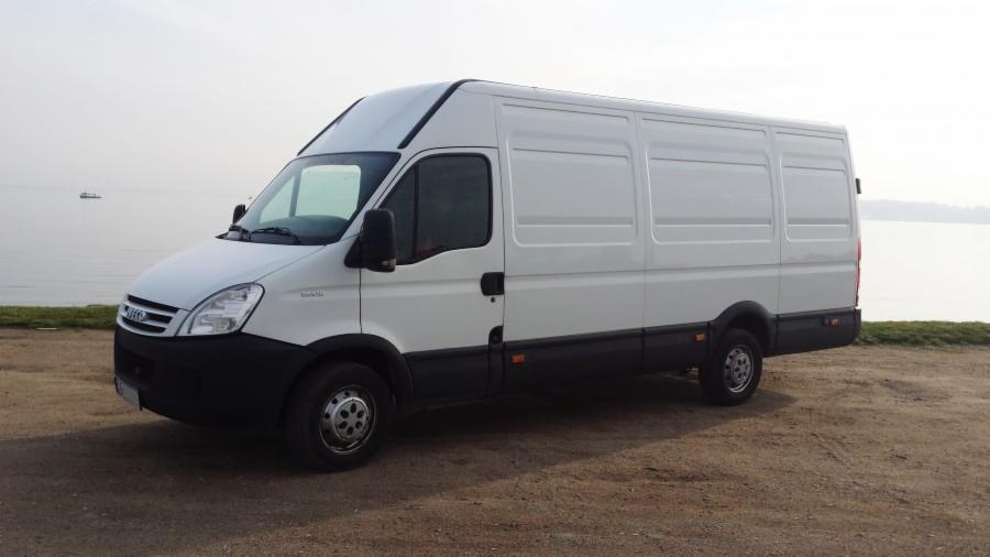 Biały bus dostawczy Iveco na tle wody i zieleni, z widocznym statkiem w oddali. Jasne niebo, spokojna woda.