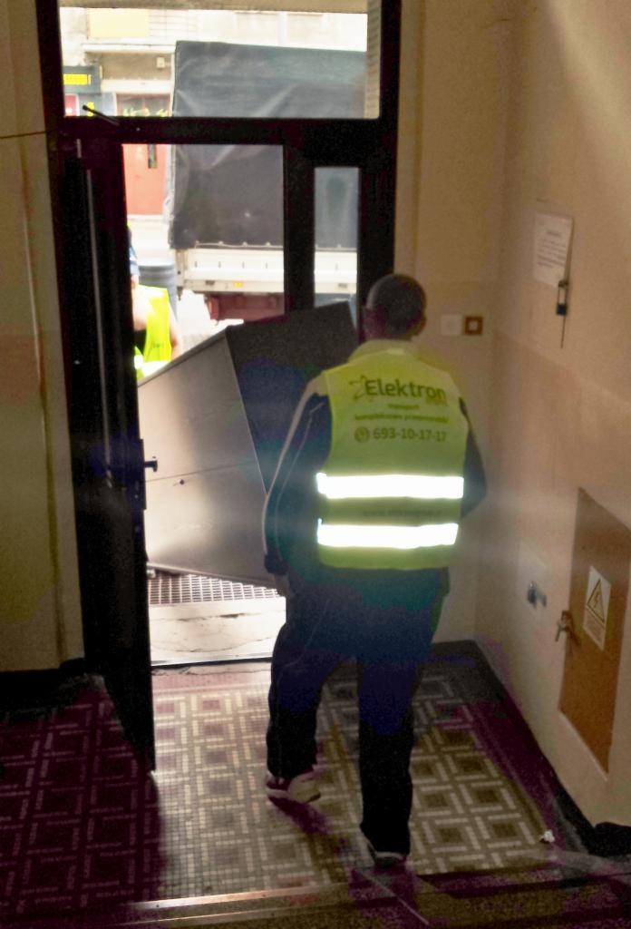 Pracownik w kamizelce firmy Elektron wnosi duży metalowy przedmiot do budynku, widoczny samochód dostawczy, przeprowadzka.