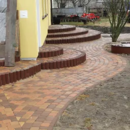 Kaskadowe schody i wijąca się ścieżka z kostki brukowej w odcieniach brązu i pomarańczy, obrzeża z brązowych palisad. Tło: żółty budynek, traktor, drzewo.