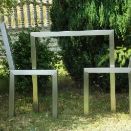 Dwa minimalistyczne, metalowe krzesła połączone ramą, stoją na trawie na tle zieleni. Krzesła wykonane z profili o kwadratowym przekroju, o surowym, nowoczesnym wyglądzie.