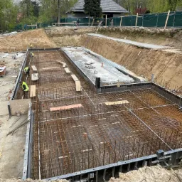 Szeroki kadr na plac budowy z wykopami i zbrojeniem fundamentów pod budynek, widoczni pracownicy w odblaskowych kamizelkach, w tle zieleń drzew i budynek z ciemnym dachem.