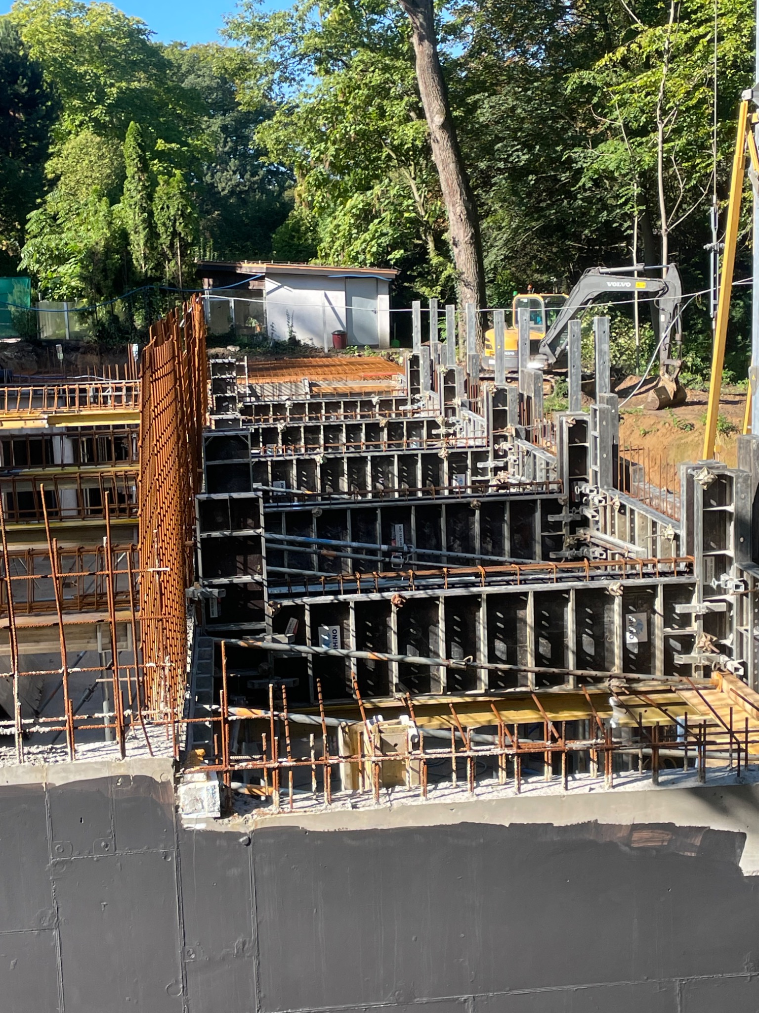 Skomplikowane, wielopoziomowe szalunki zbrojeniowe na placu budowy, z widocznym wykopem i częściowo zalanym betonem, w tle zieleń i maszyna budowlana.