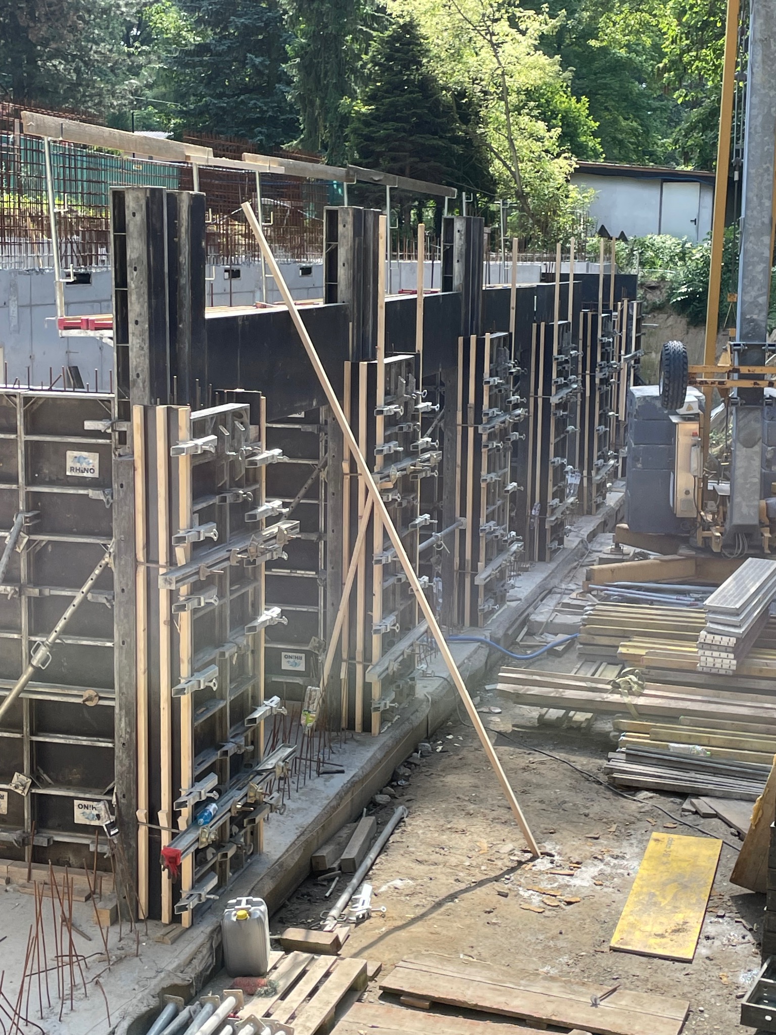 Szczegółowy widok szalunków pod fundamenty z widocznymi elementami mocującymi, zbrojeniem w tle i drewnianą podporą, na placu budowy w słoneczny dzień.