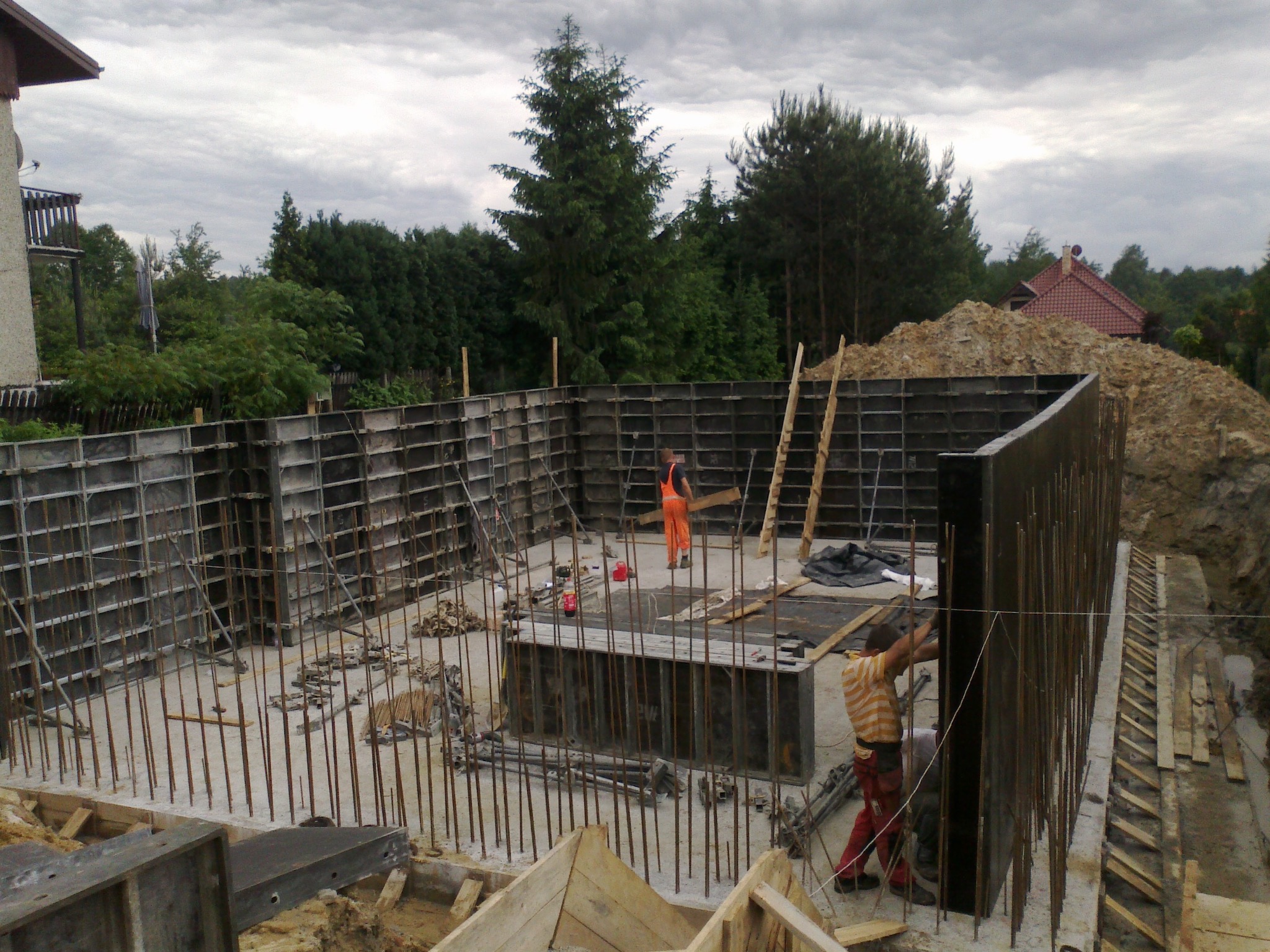 Budowa fundamentów domu z użyciem szalunków systemowych; w tle widoczny pracownik w pomarańczowym kombinezonie oraz stos drewna i wykopana ziemia.