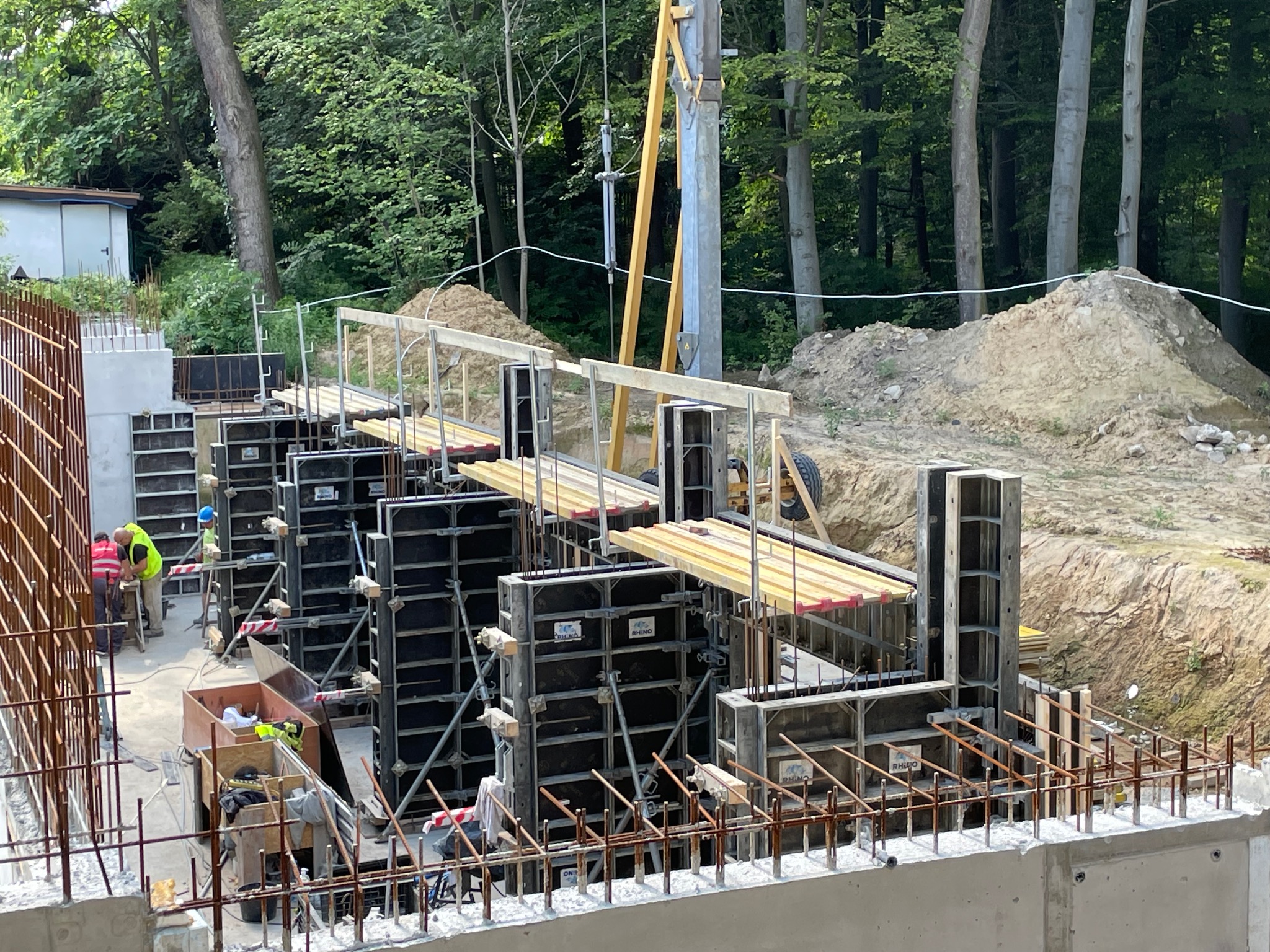 Budowa fundamentów: szalunki zbrojone stalą wystającą z betonu, pracownicy w odblaskowych kamizelkach i kaskach, w tle drzewa i mały budynek.
