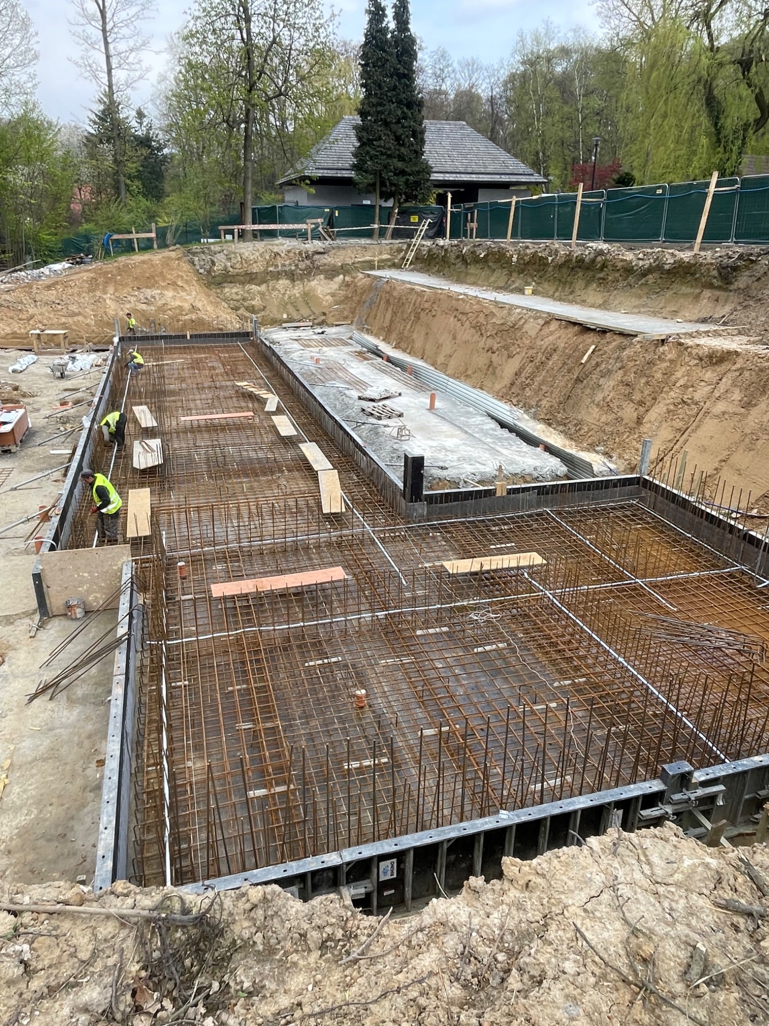 Szeroki kadr na plac budowy z wykopami i zbrojeniem fundamentów pod budynek, widoczni pracownicy w odblaskowych kamizelkach, w tle zieleń drzew i budynek z ciemnym dachem.
