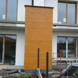 Fragment elewacji domu w trakcie wykończenia, z widoczną teksturą imitującą drewno, jasna kolorystyka, w tle okna i rusztowanie.