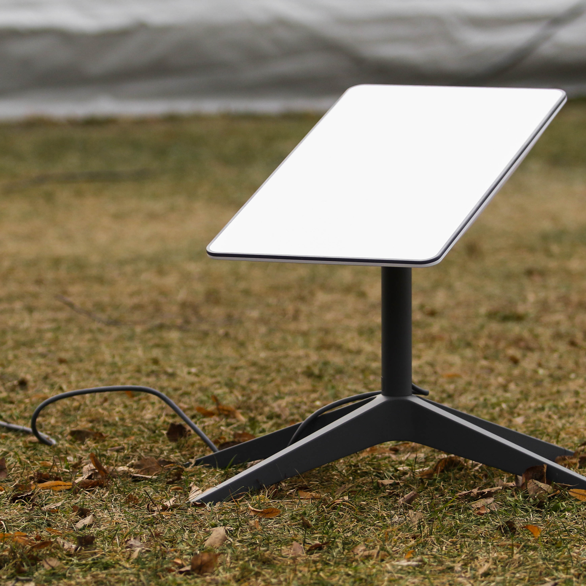 Antena Starlink na trawie, z widocznym kablem. Biała powierzchnia anteny skierowana w górę, szara podstawa. Tło rozmyte, zielono-brązowe. Nietypowe ujęcie.