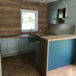 T.J.MEBLE - Jasna kuchnia z drewnianymi akcentami: fronty szafek w odcieniu błękitu, drewniana ściana nad blatem, wyspa z niebieskim panelem, podłoga ułożona w jodełkę, widoczne kable zwisające z sufitu.