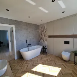 Nowoczesna łazienka z wolnostojącą wanną, umywalką nablatową, toaletą i zabudową meblową w odcieniach szarości, z drewnianą podłogą ułożoną w jodełkę i designerską lampą wiszącą.