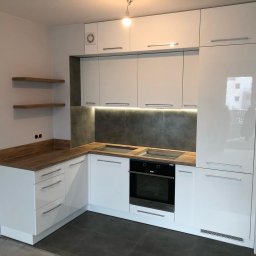 T.J.MEBLE - Nowoczesna, biała kuchnia z drewnianymi blatami, szarym backsplashiem, oświetleniem LED podszafkowym i dwoma drewnianymi półkami na ścianie. Widoczny piekarnik i płyta grzewcza. Stan surowy...