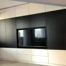 Nowoczesna zabudowa ściany z szafkami w kolorze czarnym i białym, z wnęką na telewizor. Widoczny fragment grzejnika pod oknem.