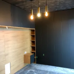 Nowoczesna sypialnia z zabudową: antracytowe szafy wnękowe na całej ścianie, drewniane panele za łóżkiem z szafką nocną i otwartymi półkami, trzy złote lampy wiszące na tle szarego sufitu.