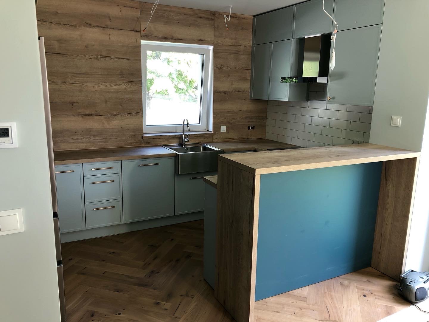 Jasna kuchnia z drewnianymi akcentami: fronty szafek w odcieniu błękitu, drewniana ściana nad blatem, wyspa z niebieskim panelem, podłoga ułożona w jodełkę, widoczne kable zwisające z sufitu.