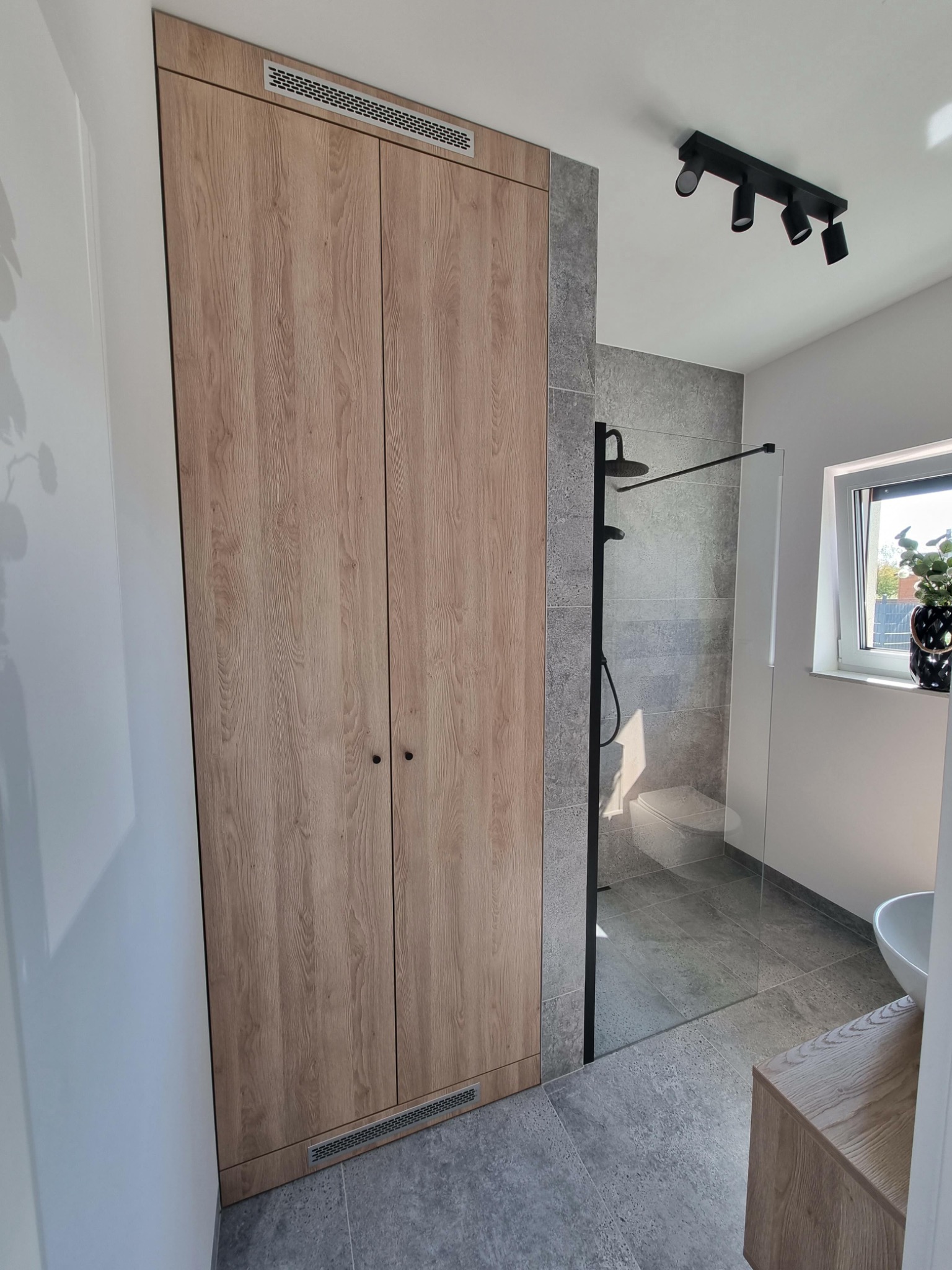 Wysoka, drewniana szafa z wentylacją wtopiona w ścianę łazienki, obok przeszklony prysznic z czarną armaturą i szarą okładziną, widoczna umywalka i szafka pod umywalką.